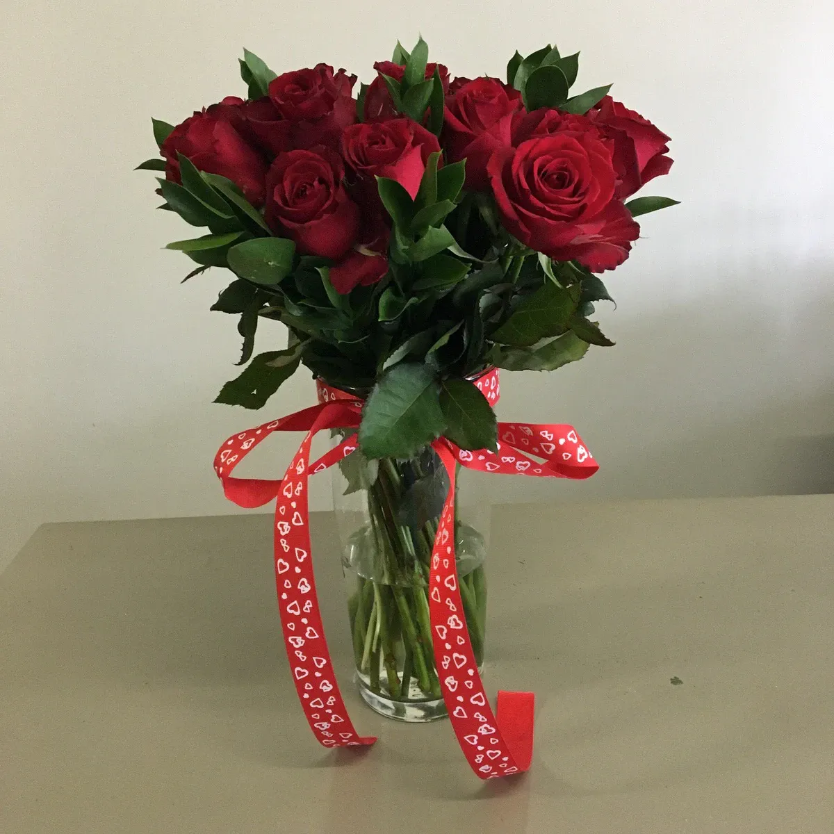 Ten red roses with greenery in a glass vase tied with a heart-print ribbon