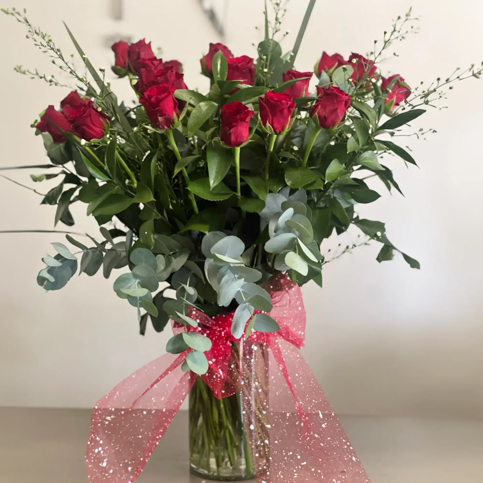 Twenty long-stemmed red roses with eucalyptus in a glass vase with a red organza bow
