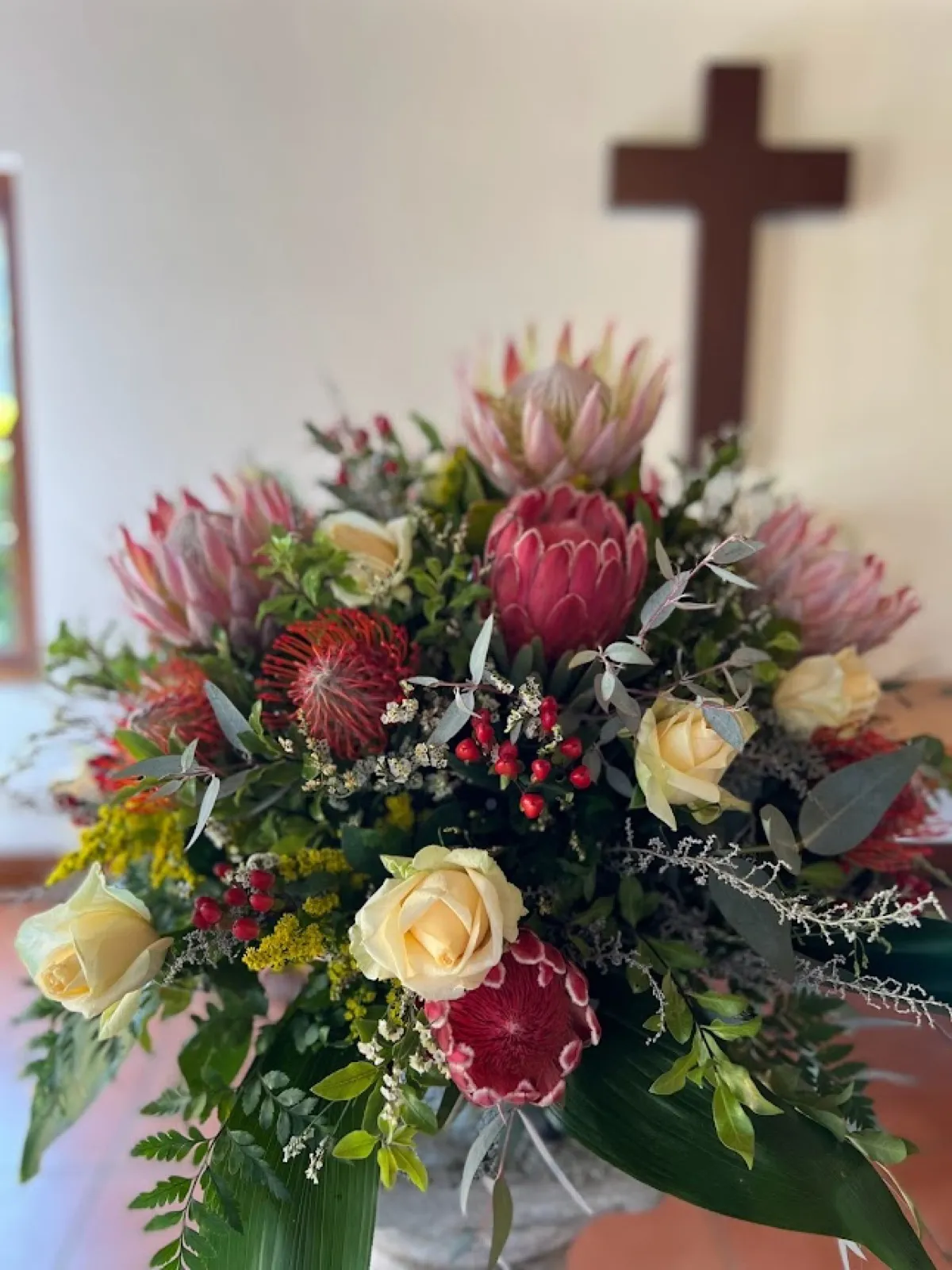 Church arrangement of pink king proteas, pincushions and cream roses in front of a wooden cross