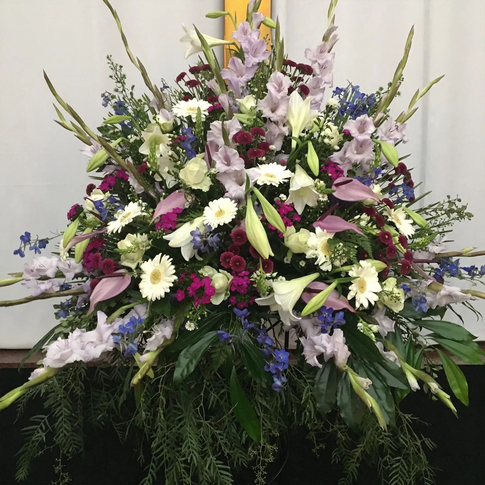 Large fan-shaped church arrangement of lilac gladioli, white lilies, gerberas and blue delphiniums