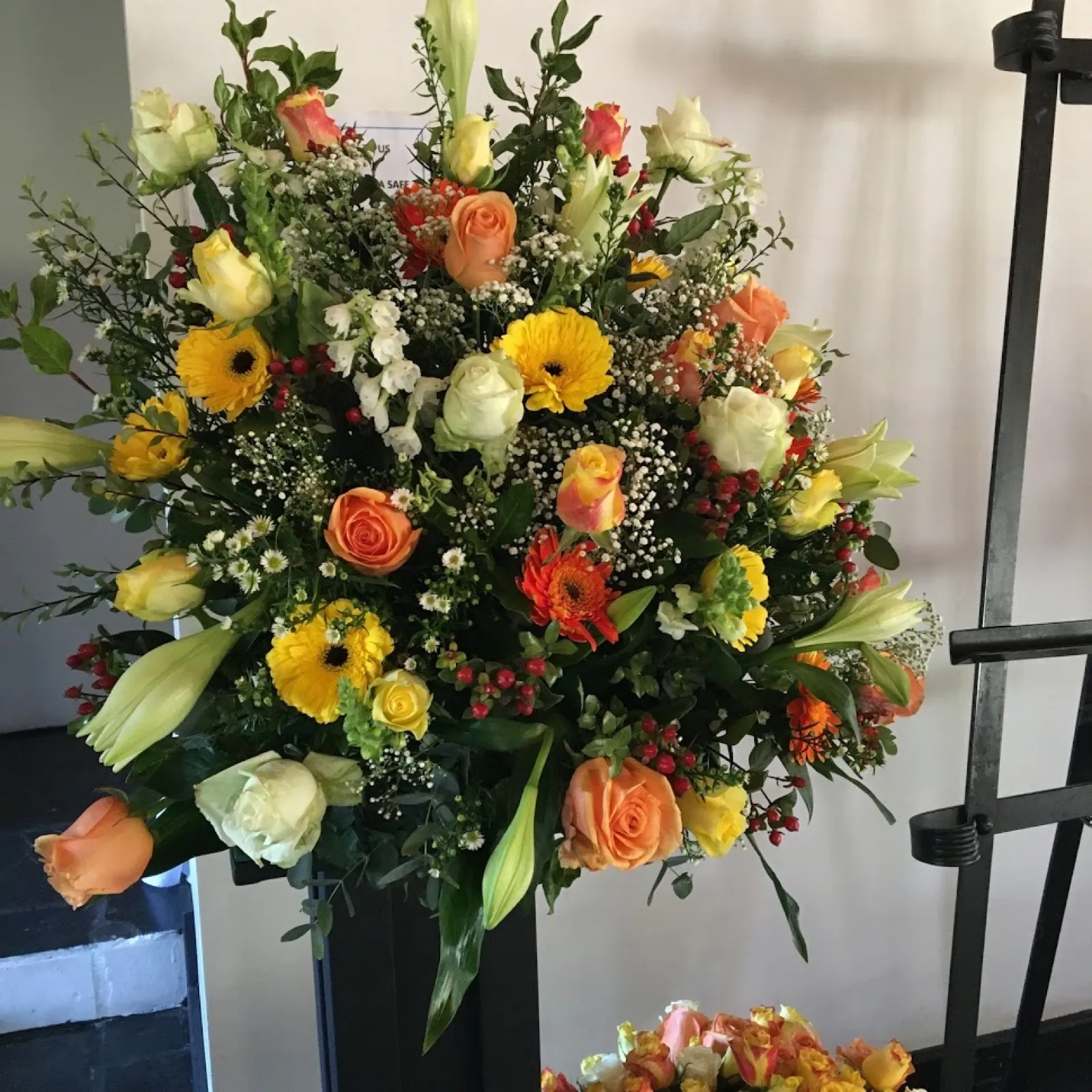 Sunny church arrangement of yellow gerberas, peach roses and lilies on a black pedestal