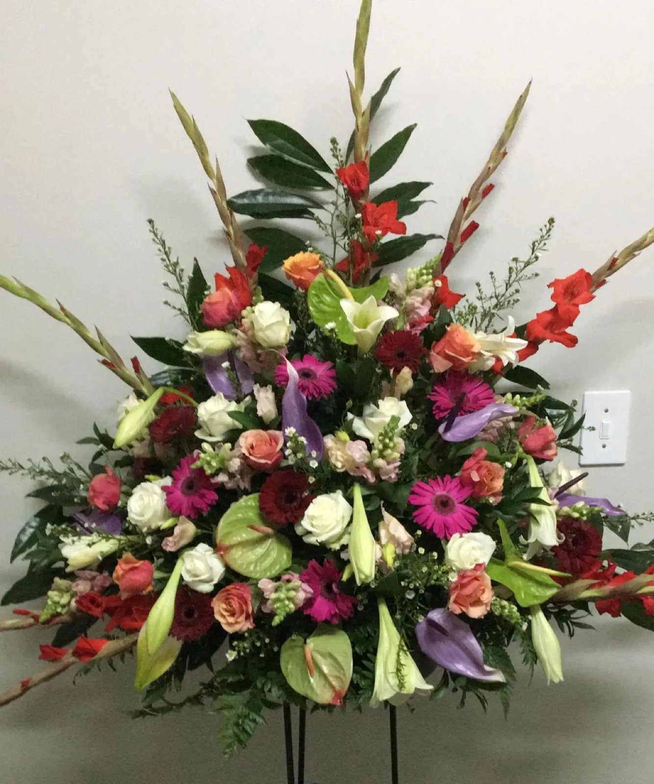 Bright fan-shaped spray of red gladioli, pink gerberas, roses and anthuriums on a stand