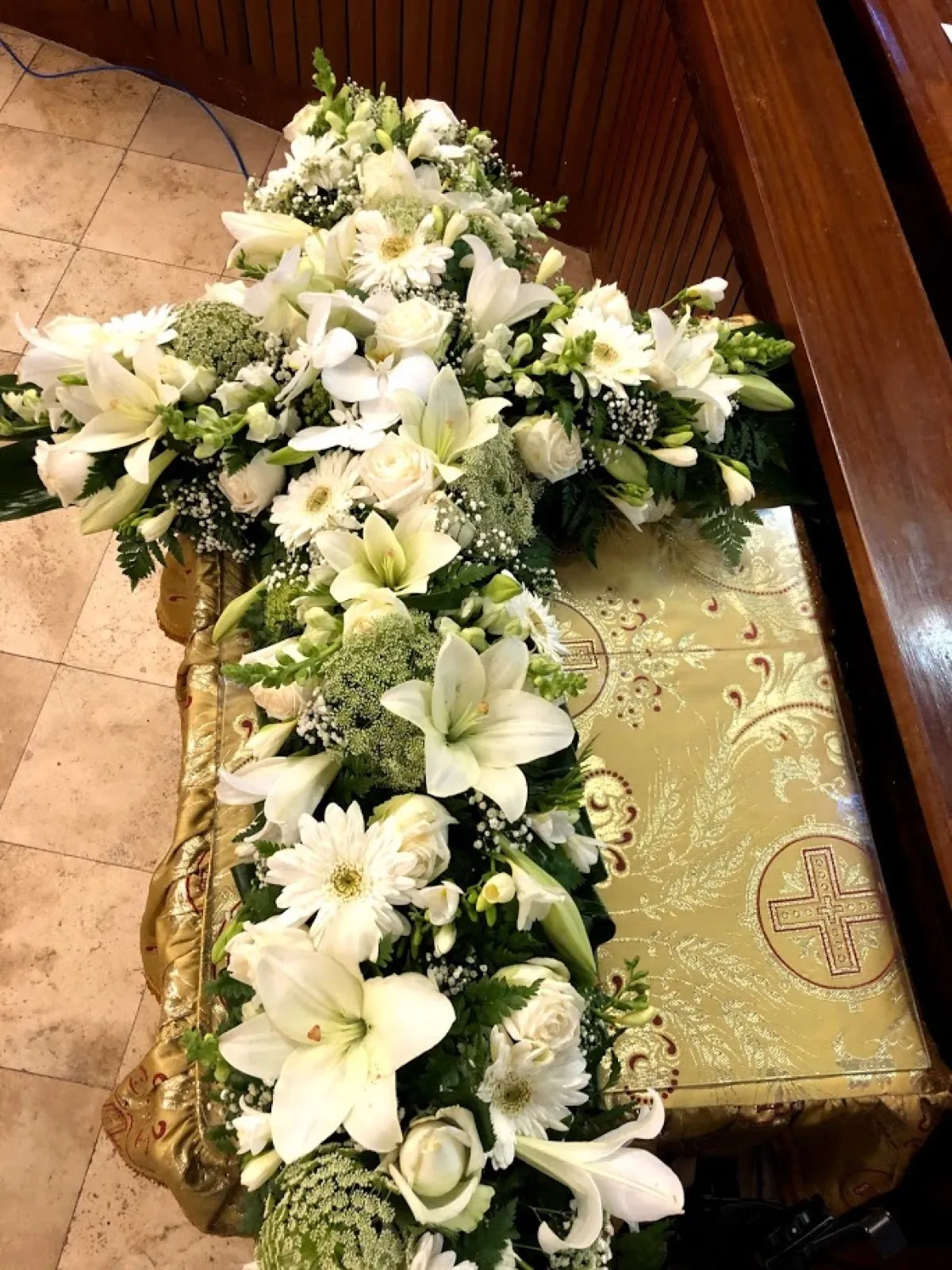 White flower cross of lilies, roses and gerberas laid on a gold-draped coffin in a church