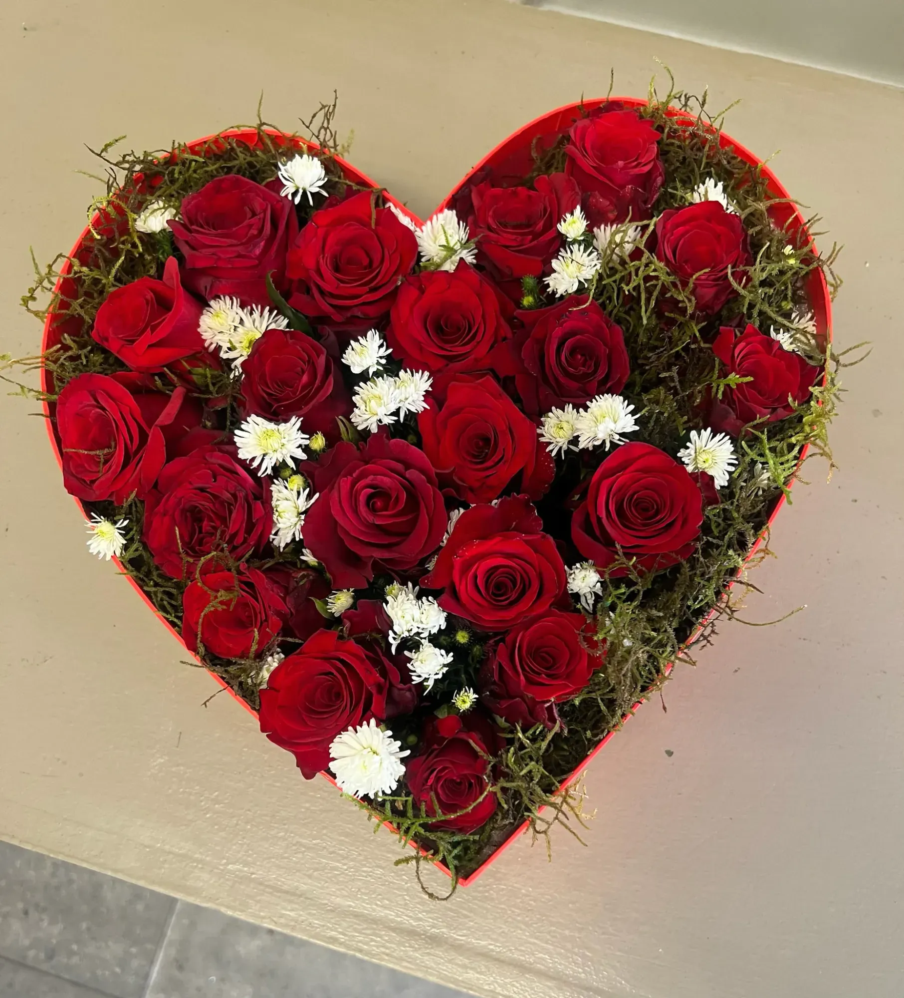 Heart-shaped red box filled with red roses and small white daisies, seen from above