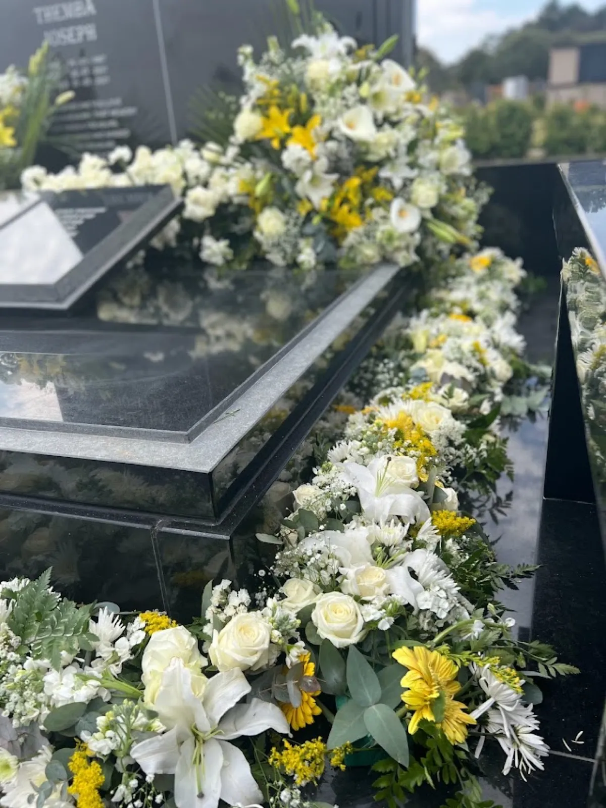 Garland of white roses, lilies and yellow chrysanthemums along a black granite headstone