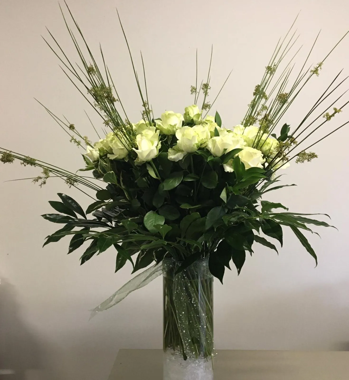 White roses with tall grasses and dark foliage in a tall glass cylinder vase